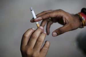 A man passes a cigarette to another while smoking as they sit on a pavement along a road in New Delhi